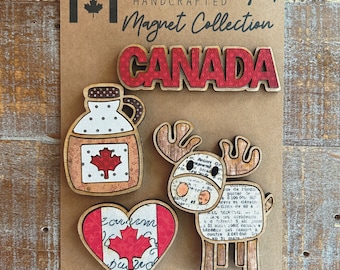 May include: A set of four wooden magnets featuring Canadian themes. The magnets are shaped like a maple leaf, a moose, a jug of maple syrup, and the word "CANADA". The magnets are painted in red, white, and brown colours and have a rustic, handcrafted look.