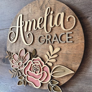 May include: A round wooden sign with the name "Amelia Grace" written in gold script. The sign is decorated with pink and brown flowers and leaves.