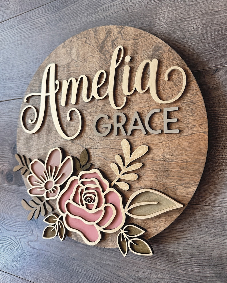 May include: A round wooden sign with the name "Amelia Grace" written in gold script. The sign is decorated with pink and brown flowers and leaves.