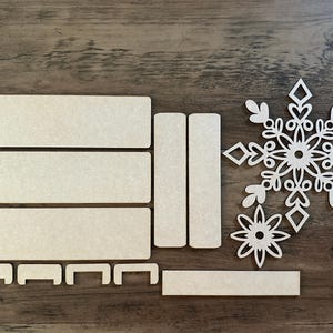 Puede incluir: Un conjunto de piezas artesanales de madera para crear un adorno de copo de nieve. El conjunto incluye un dise&ntilde;o de copo de nieve, una estrella y varias piezas rectangulares.