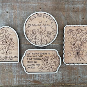 May include: Four wooden decorative signs with floral designs and white borders. One sign is arched with the text "WHERE FLOWERS BLOOM, SO DOES HOPE." Another is round with "Know that you are beautiful." A third is rectangular with "DON'T WAIT FOR SOMEONE TO BRING YOU FLOWERS. PLANT YOUR OWN GARDEN AND DECORATE YOUR OWN SOUL."