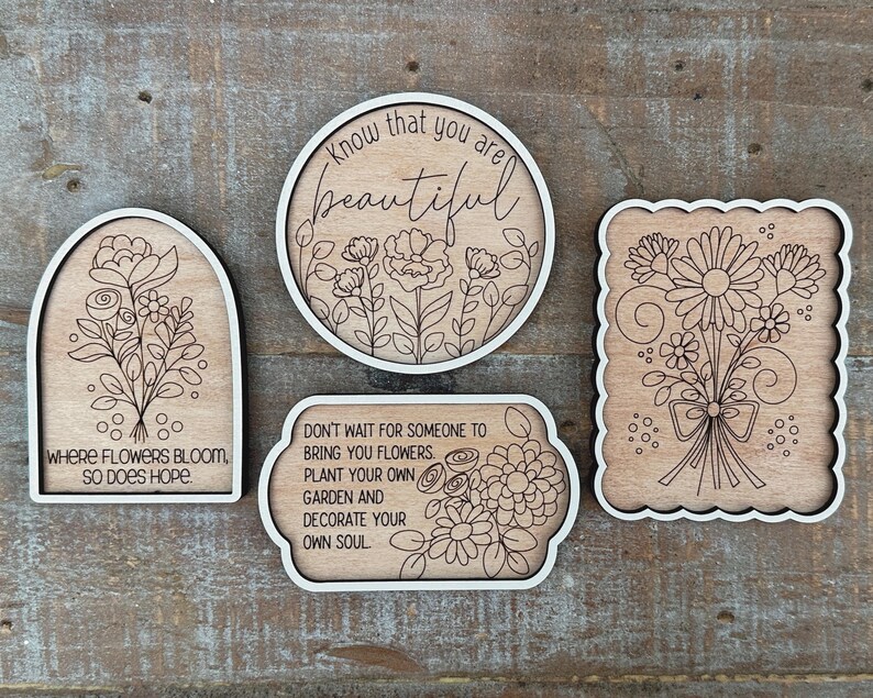 May include: Four wooden decorative signs with floral designs and white borders. One sign is arched with the text "WHERE FLOWERS BLOOM, SO DOES HOPE." Another is round with "Know that you are beautiful." A third is rectangular with "DON'T WAIT FOR SOMEONE TO BRING YOU FLOWERS. PLANT YOUR OWN GARDEN AND DECORATE YOUR OWN SOUL."