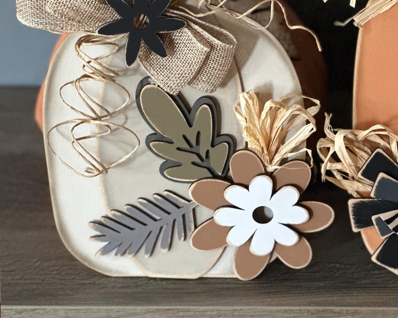 Pode incluir: Uma ab&oacute;bora de madeira branca decorada com um la&ccedil;o de juta marrom, flores pretas, folhas marrons e verdes e flores marrons e brancas. A ab&oacute;bora &eacute; decorada com barbante marrom e branco.