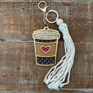 May include: A wooden coffee cup keychain with a black and white polka dot pattern, a red heart, and a gold trim. The keychain has a white tassel attached to a silver key ring.