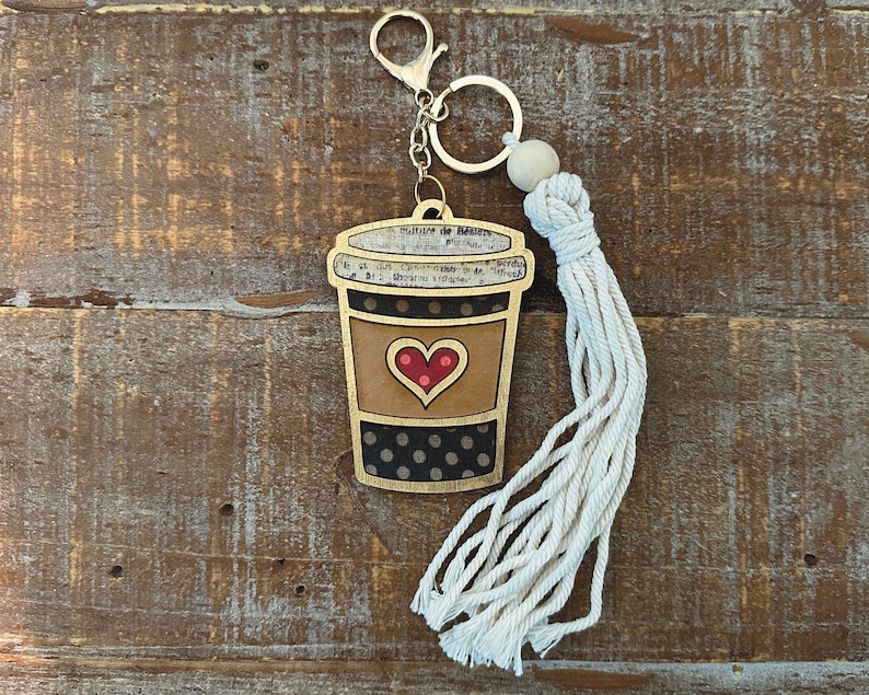 May include: A wooden coffee cup keychain with a black and white polka dot pattern, a red heart, and a gold trim. The keychain has a white tassel attached to a silver key ring.