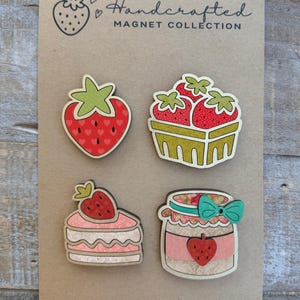 May include: A collection of handcrafted strawberry magnets on a brown card. The magnets include a whole strawberry, a basket of strawberries, a slice of cake with a strawberry on top, and a jar of strawberry jam. The text "Handcrafted MAGNET COLLECTION" is at the top.