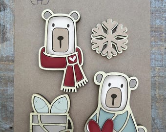 May include: Set of five wooden magnets featuring a winter theme. The magnets include a polar bear wearing a red scarf, a polar bear holding a red heart, a polar bear wearing a blue scarf, a snowflake, and a bucket with a lid.