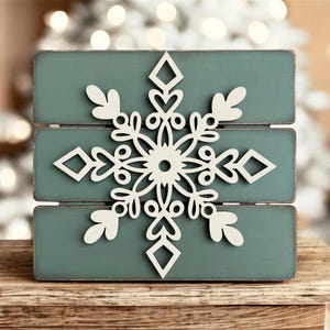 Puede incluir: Un letrero de madera de color verde azulado con un dise&ntilde;o de copo de nieve recortado en blanco. El letrero est&aacute; sobre una superficie de madera.