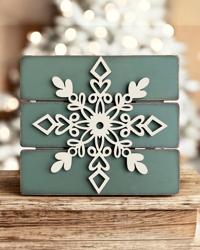 Puede incluir: Un letrero de madera de color verde azulado con un dise&ntilde;o de copo de nieve recortado en blanco. El letrero est&aacute; sobre una superficie de madera.