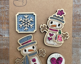 May include: Set of four handcrafted wooden magnets featuring a snowflake, a heart, and two snowmen. The snowmen are decorated with pink and blue fabric scraps and have a vintage look. The magnets are part of a "Handcrafted Magnet Collection".
