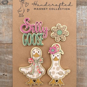 May include: A collection of handcrafted magnets on a brown card. The magnets include a flower, the words "Silly Goose", and two goose figures with floral accents. The card also features a goose illustration and the text "Handcrafted Magnet Collection."