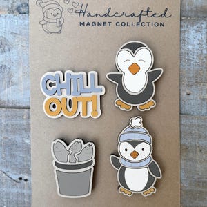 Puede incluir: Un conjunto de tres imanes de madera sobre una tarjeta marrón etiquetada Handcrafted Magnet Collection. Los imanes incluyen el texto CHILL OUT! en azul y amarillo, un pingüino con vientre blanco y pico naranja, y un cubo de tres peces con pingüino.