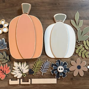 Pode incluir: Um conjunto de recortes de madeira de ab&oacute;bora e folhas para artesanato de outono. O conjunto inclui duas ab&oacute;boras, uma laranja e uma branca, juntamente com v&aacute;rias folhas em tons de verde, castanho e vermelho. Tamb&eacute;m existem v&aacute;rios recortes de flores em branco, castanho e preto.