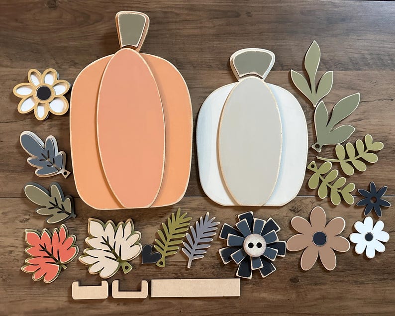 Pode incluir: Um conjunto de recortes de madeira de ab&oacute;bora e folhas para artesanato de outono. O conjunto inclui duas ab&oacute;boras, uma laranja e uma branca, juntamente com v&aacute;rias folhas em tons de verde, castanho e vermelho. Tamb&eacute;m existem v&aacute;rios recortes de flores em branco, castanho e preto.