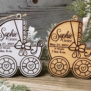 May include: Two wooden baby carriage ornaments with the name "Sophie Grace" and the date "May 21, 2021" and the time "3:18 pm" and the weight "7 lbs. 6 oz.". One ornament is painted white and the other is natural wood.
