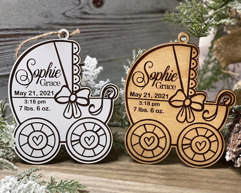 May include: Two wooden baby carriage ornaments with the name "Sophie Grace" and the date "May 21, 2021" and the time "3:18 pm" and the weight "7 lbs. 6 oz.". One ornament is painted white and the other is natural wood.
