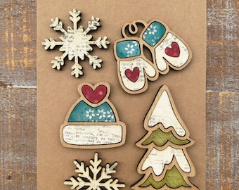 May include: Set of five handcrafted wooden magnets with a winter theme. The magnets feature a snowflake, a pair of mittens, a winter hat, a pine tree, and another snowflake. The magnets are decorated with a vintage paper design and have red hearts.