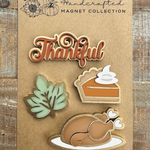 May include: A set of four Thanksgiving-themed magnets featuring a turkey, a pie, a leaf, and the word "Thankful". The magnets are made of wood and have a rustic, handcrafted look.