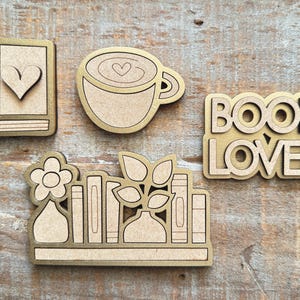 May include: A collection of wooden cutouts with a gold border. The set includes a book with a heart, a coffee cup with a heart, the words "BOOK LOVER", and a shelf with books, a flower, and leaves. The items are on a wooden surface.