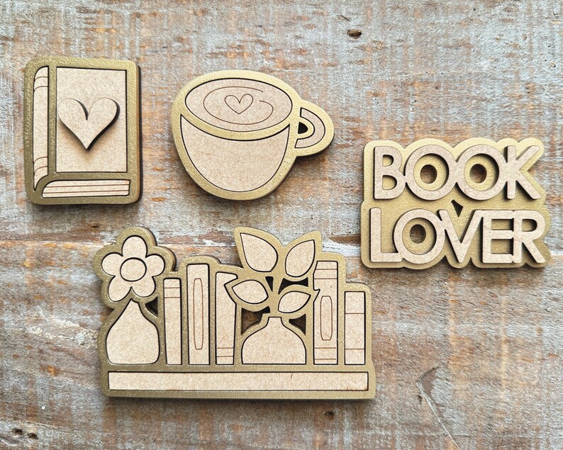 May include: A collection of wooden cutouts with a gold border. The set includes a book with a heart, a coffee cup with a heart, the words "BOOK LOVER", and a shelf with books, a flower, and leaves. The items are on a wooden surface.