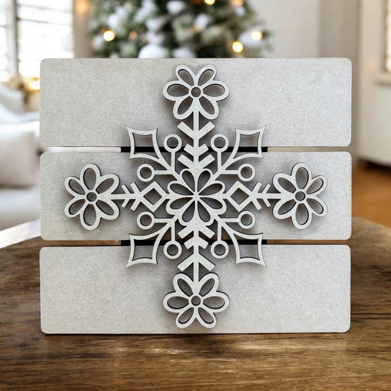 Puede incluir: Un adorno de copo de nieve de madera blanca con un dise&ntilde;o decorativo. El copo de nieve est&aacute; unido a una base de madera blanca.