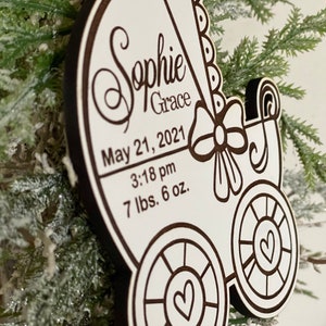 May include: White wooden baby carriage ornament with a black outline. The ornament is personalized with the name "Sophie Grace", the date "May 21, 2021", the time "3:18 pm", and the weight "7 lbs. 6 oz."