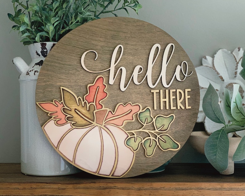 May include: A round wooden sign with the words 'hello there' in white script. The sign is decorated with a pink and green pumpkin and leaves.