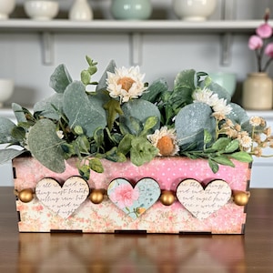 May include: A rectangular wooden floral arrangement box with a pink floral pattern and three heart-shaped embellishments. The box is filled with greenery and white and orange flowers. Gold-colored spheres are placed between the hearts.