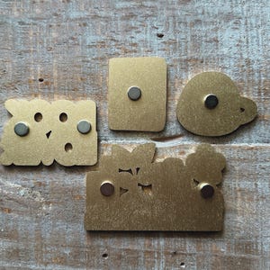 May include: Four brass-colored metal magnets in various shapes: a rectangle, a rounded shape, and two with cut-out designs. Each magnet has a small, round, dark gray magnet attached to the back. The magnets are displayed on a weathered, light-colored wooden surface.