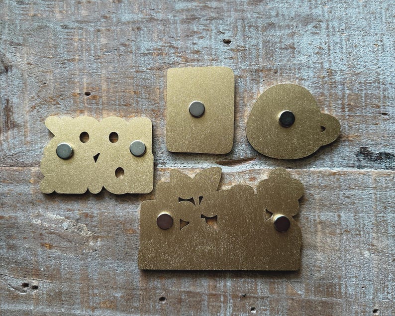 May include: Four brass-colored metal magnets in various shapes: a rectangle, a rounded shape, and two with cut-out designs. Each magnet has a small, round, dark gray magnet attached to the back. The magnets are displayed on a weathered, light-colored wooden surface.