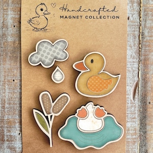 May include: A set of five wooden magnets featuring a duck theme. The magnets include a duck in a pond, a duckling, a cloud with raindrops, a flower, and a duck with a patterned design. The magnets are handcrafted and come in a variety of colors, including blue, green, yellow, orange, and brown. "Handcrafted Magnet Collection" is printed on the card.