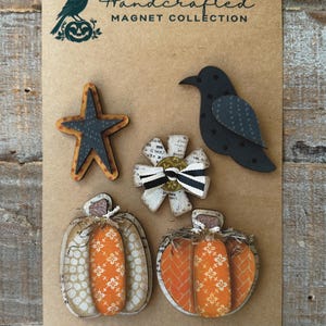 May include: A collection of handcrafted magnets on a brown card. The magnets include a star, a crow, a flower, and two pumpkins. The pumpkins are orange with white floral patterns. The card reads "Handcrafted Magnet Collection".