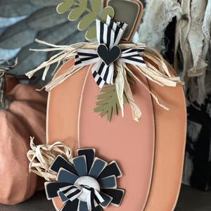 Pode incluir: Uma ab&oacute;bora de madeira cor de p&ecirc;ssego com uma fita listrada branca e preta, um cora&ccedil;&atilde;o preto e uma flor preta e cinza. A ab&oacute;bora &eacute; decorada com palha e folhas verdes.