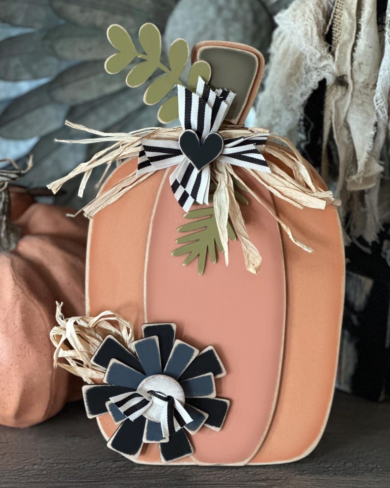 Pode incluir: Uma ab&oacute;bora de madeira cor de p&ecirc;ssego com uma fita listrada branca e preta, um cora&ccedil;&atilde;o preto e uma flor preta e cinza. A ab&oacute;bora &eacute; decorada com palha e folhas verdes.