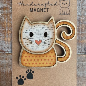 May include: Handcrafted wooden cat magnet with a brown and white polka dot pattern. The cat has a pink heart on its chest and black paw prints are on the bottom of the magnet. The text "Handcrafted Magnet" is on the top of the magnet.