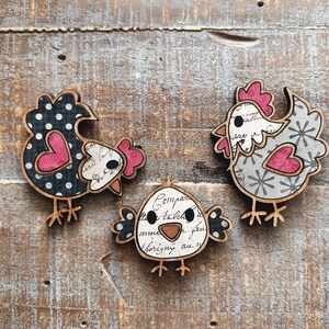 Puede incluir: Tres adornos de pollo de madera con un dise&ntilde;o de estilo vintage. Los pollos est&aacute;n decorados con lunares negros y blancos, patrones florales grises y blancos y letras negras sobre fondo blanco. Cada pollo tiene un coraz&oacute;n rosa en el pecho.