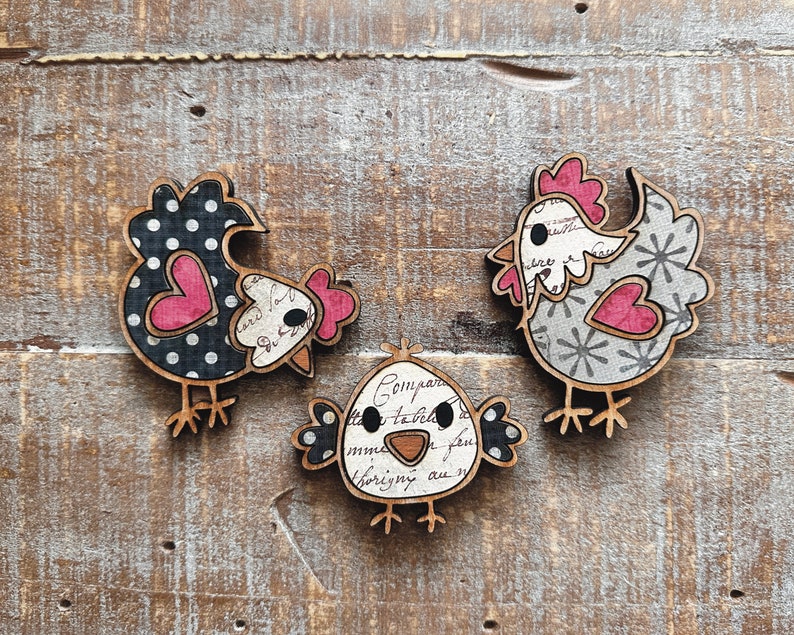 Puede incluir: Tres adornos de pollo de madera con un dise&ntilde;o de estilo vintage. Los pollos est&aacute;n decorados con lunares negros y blancos, patrones florales grises y blancos y letras negras sobre fondo blanco. Cada pollo tiene un coraz&oacute;n rosa en el pecho.