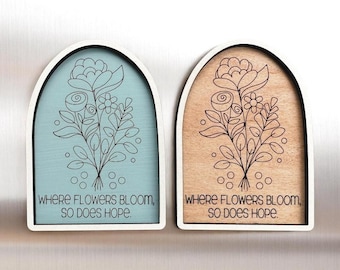 May include: Two wooden wall art pieces with arched tops. One is light blue, the other is tan. Both feature a black floral design and the text "WHERE FLOWERS BLOOM, SO DOES HOPE."