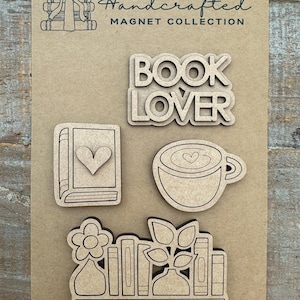 May include: A collection of handcrafted wooden magnets on a tan background. The magnets include a stack of books with a bow, the words "BOOK LOVER", a book with a heart, a coffee cup with a heart, and a shelf with books, plants, and a flower.