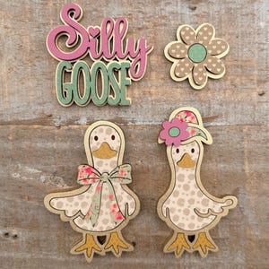 May include: Wooden cutouts of a flower, two geese, and the words "Silly Goose". The geese have floral patterned bows and hats. The flower has a heart-shaped pattern. The text is in pink and green.