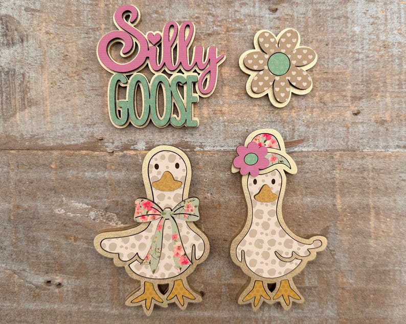 May include: Wooden cutouts of a flower, two geese, and the words "Silly Goose". The geese have floral patterned bows and hats. The flower has a heart-shaped pattern. The text is in pink and green.