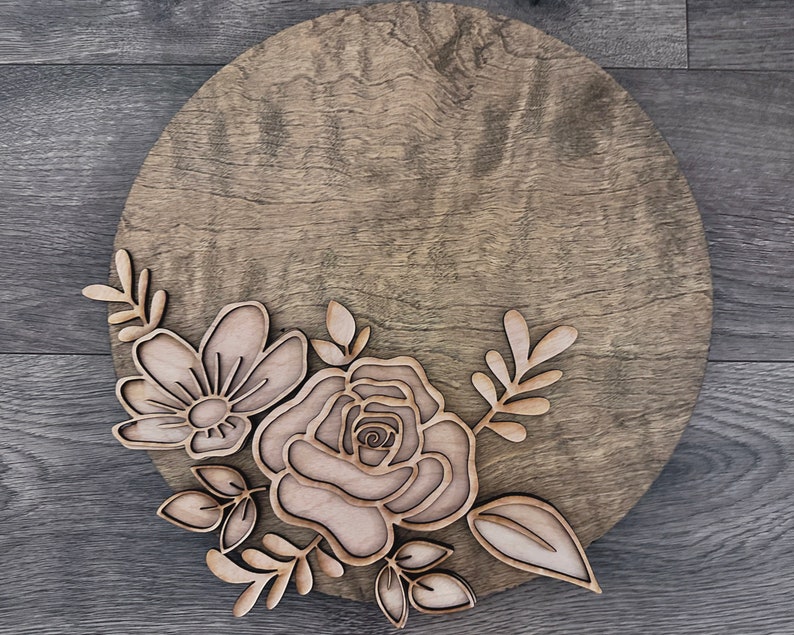 May include: A round wooden sign with a floral design. The design features a large rose and smaller flowers with leaves.