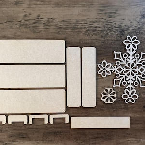 Puede incluir: Un conjunto de piezas de madera para crear un adorno de copo de nieve. El conjunto incluye cinco piezas rectangulares, dos piezas rectangulares largas y un dise&ntilde;o de copo de nieve con detalles intrincados.