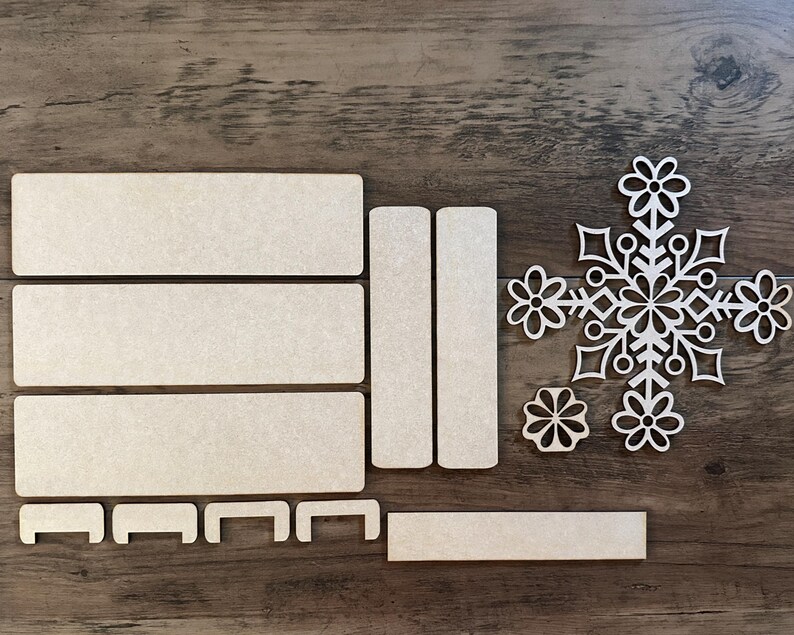 Puede incluir: Un conjunto de piezas de madera para crear un adorno de copo de nieve. El conjunto incluye cinco piezas rectangulares, dos piezas rectangulares largas y un dise&ntilde;o de copo de nieve con detalles intrincados.