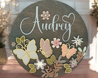May include: A round wooden sign with the name "Audrey" in white script. The sign is decorated with butterflies, flowers, and leaves in shades of pink, blue, and green.