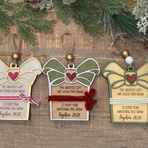 May include: Three wooden Christmas ornaments shaped like presents with a bow and ribbon. Each ornament has a different colored background and ribbon. The ornaments have the text "The greatest gift we could ever know is every year watching you grow Sophia 2021" on them.