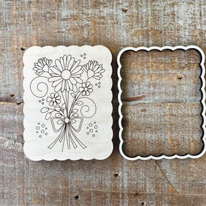 May include: A wooden cookie cutter set featuring a floral bouquet design. The set includes a scalloped-edged cutter with the floral design and a matching scalloped-edged frame. The design includes daisies, other flowers, and a bow.