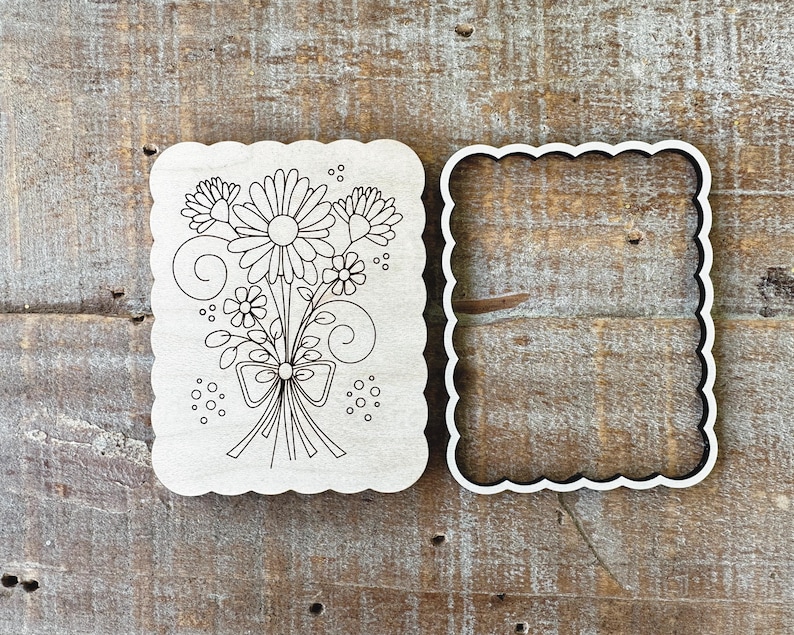 May include: A wooden cookie cutter set featuring a floral bouquet design. The set includes a scalloped-edged cutter with the floral design and a matching scalloped-edged frame. The design includes daisies, other flowers, and a bow.