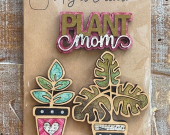 May include: A set of four wooden magnets featuring a plant theme. The magnets include a sign that says "Plant Mom" in gold letters on a pink background, a potted plant with green leaves and a pink pot with a heart, and a potted plant with green leaves and a black and white pot with polka dots.