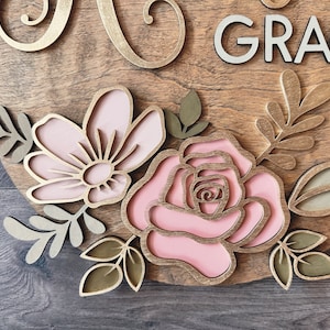 May include: A wooden sign with the name "Grace" in white letters and a floral design featuring pink and gold flowers and leaves.
